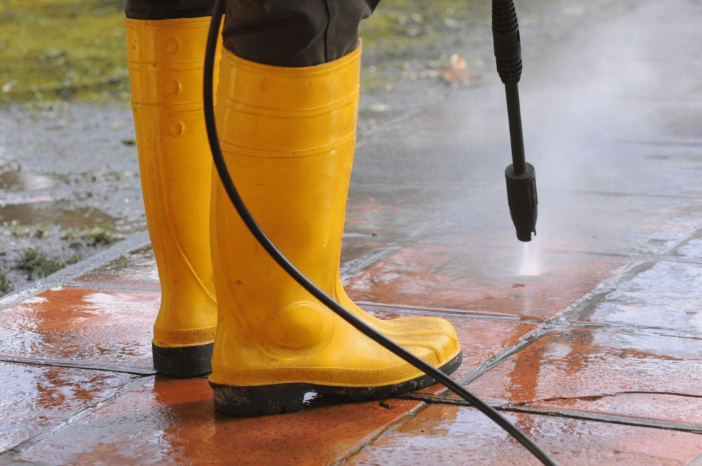 A person wearing yellow rubber boots and using a pressure washing wand to clean a surface.