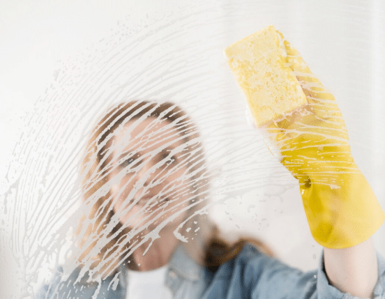 A smiling woman cleaning a glass window with a soapy sponge.